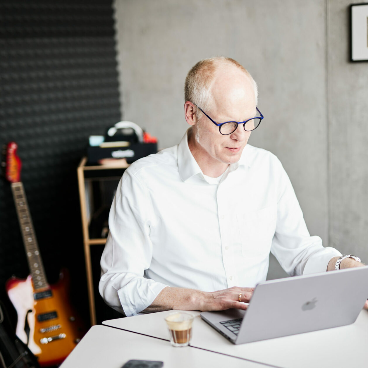 An older man working on his laptop and reaching for his smartphone – electric guitars can be seen in the background.