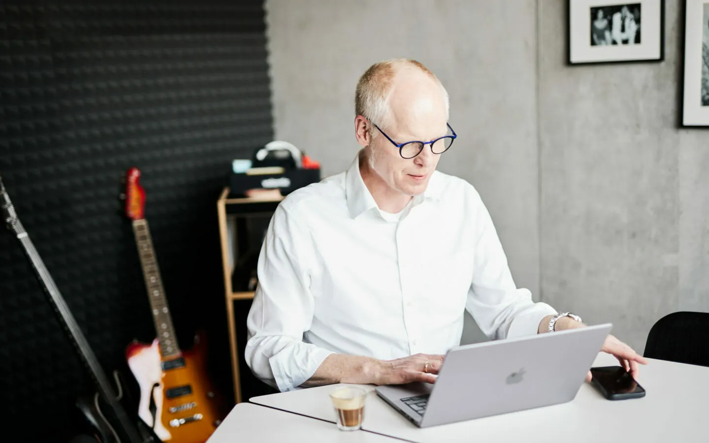 An older man working on his laptop and reaching for his smartphone – electric guitars can be seen in the background.