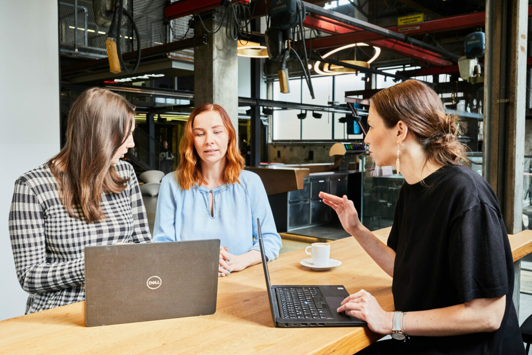 Foto von einer jungen Frau, die ihren beiden Kolleginnen eine Strategie erklärt.