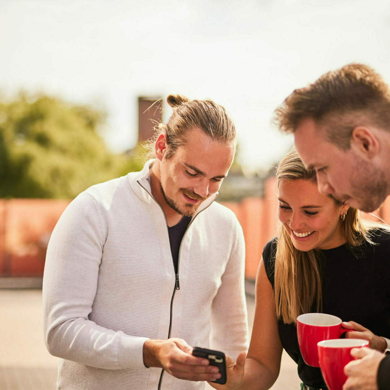 Bild von drei Kolleg*innen, die sich zusammen in der Kaffeepause etwas auf einem Smartphone ansehen.