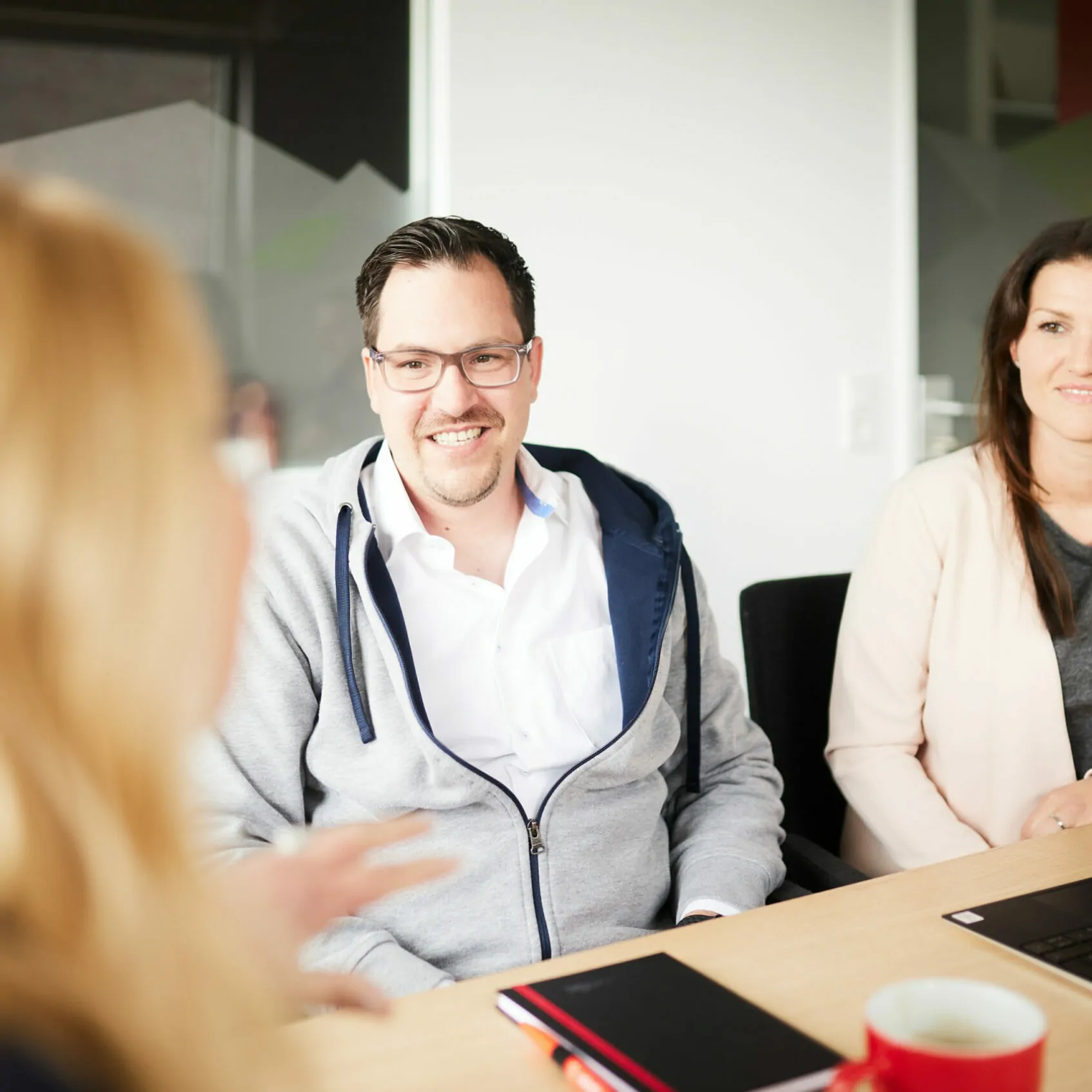 Foto von einem Mann und einer Frau, die an einem Konferenztisch sitzen und sich von einer Dame beraten lassen.