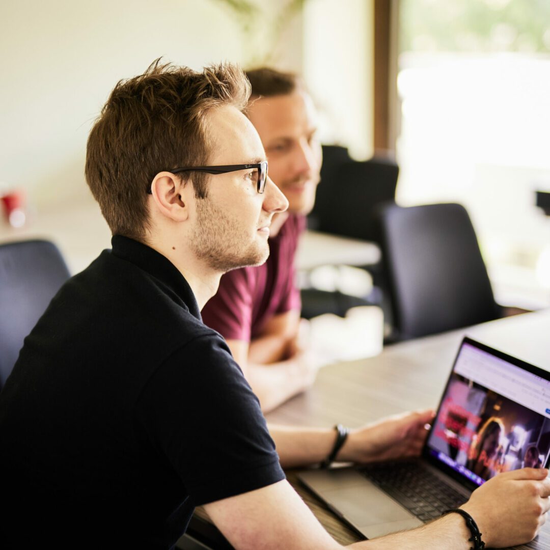 Foto von einem jungen Mann mit Brille, der an seinem Laptop sitzt und zu einem Kollegen nach vorne sieht.