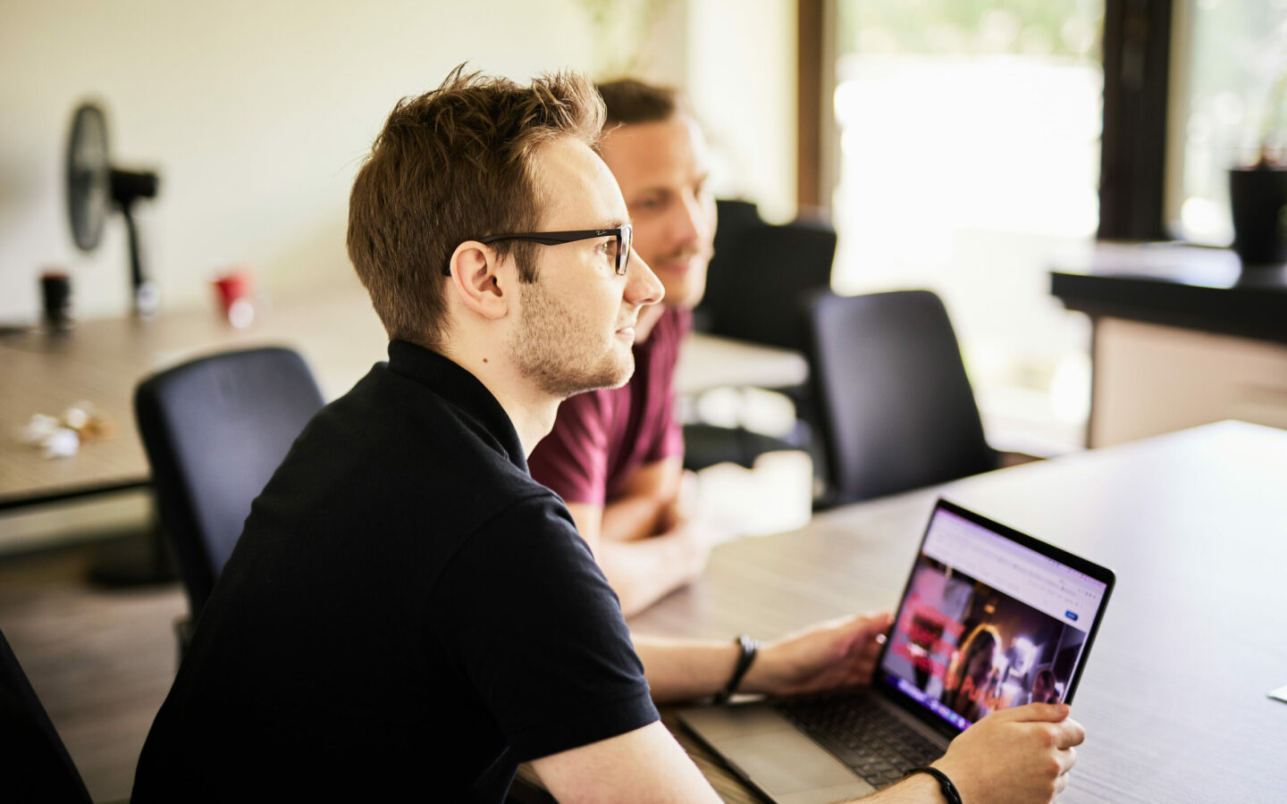 Photo of a young man with glasses sitting at his laptop and looking at a colleague.