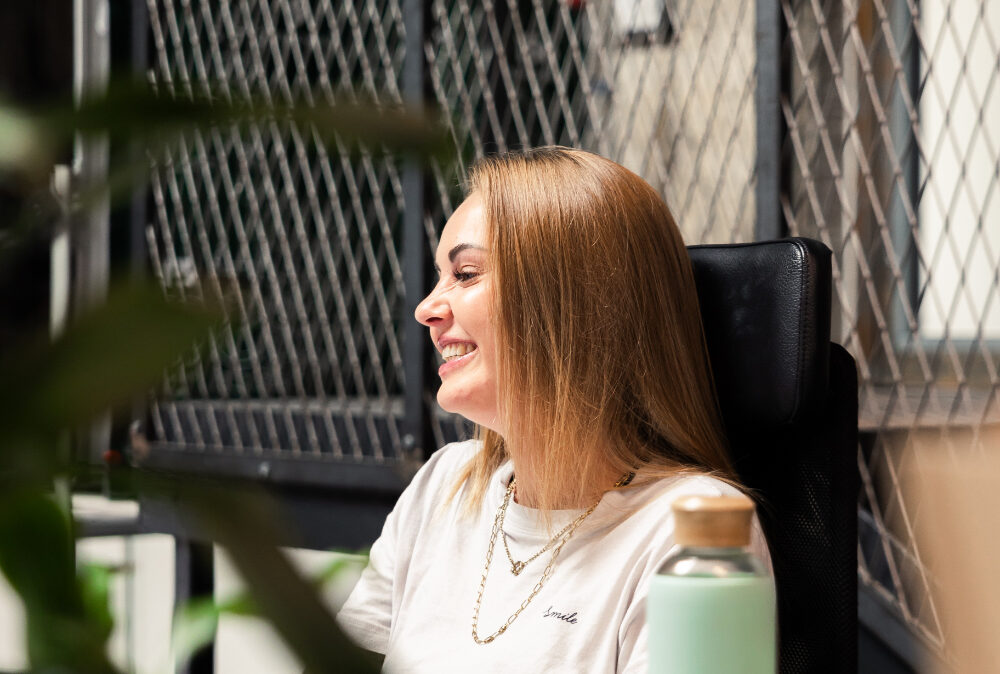 Eine Frau sitzt im Büro am Schreibtisch, lächelt und arbeitet am Computer.