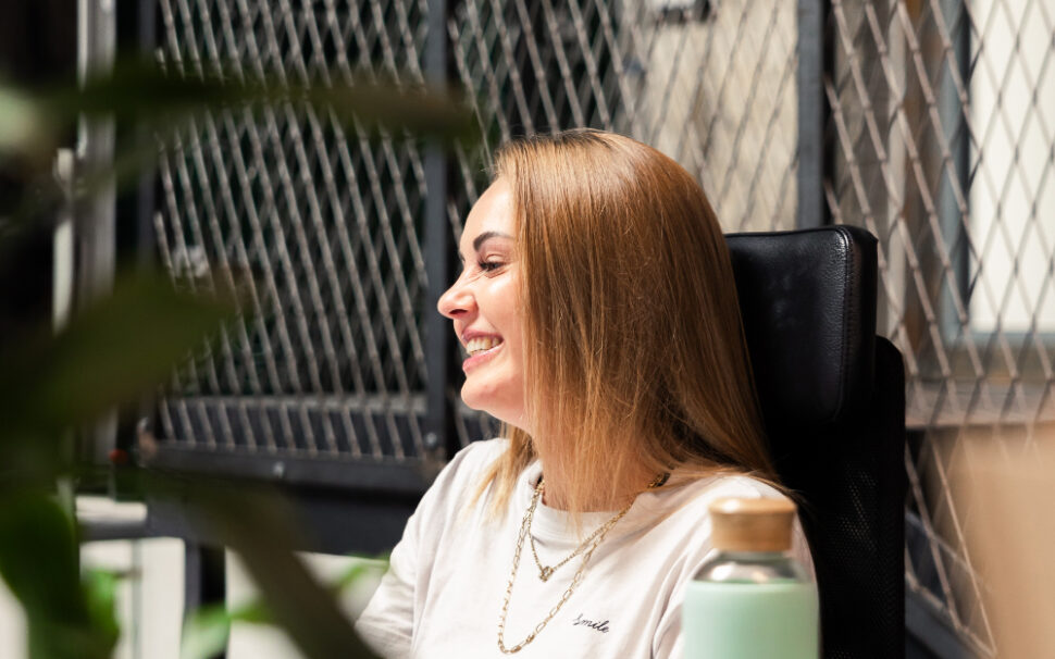 Eine Frau sitzt im Büro am Schreibtisch, lächelt und arbeitet am Computer.