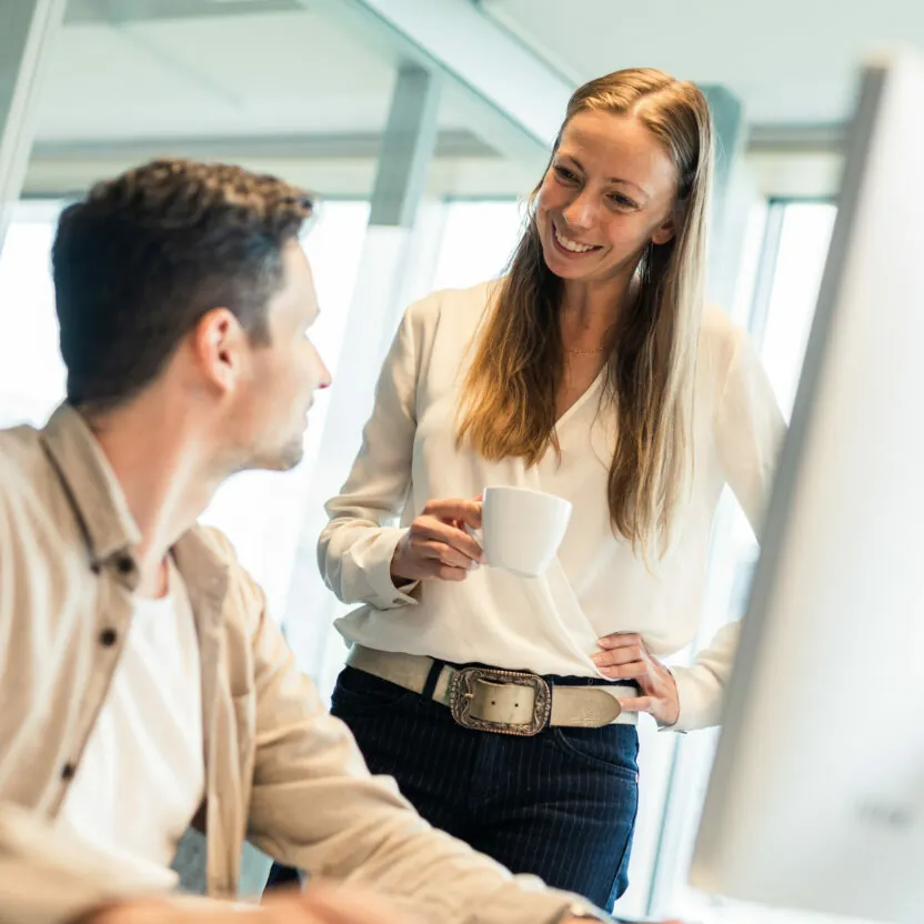 Photo of smiling woman with a coffee cup in hand explaining something to her colleague.