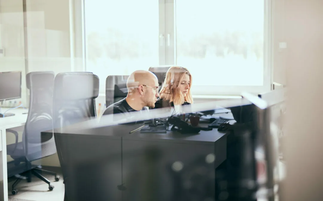 Photo of a man and a woman sitting at an office workstation, talking to each other.