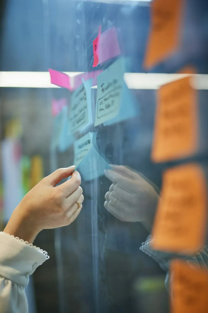 Bildausschnitt von einer Frauenhand, die ein Post-It von einer Glaswand nimmt.