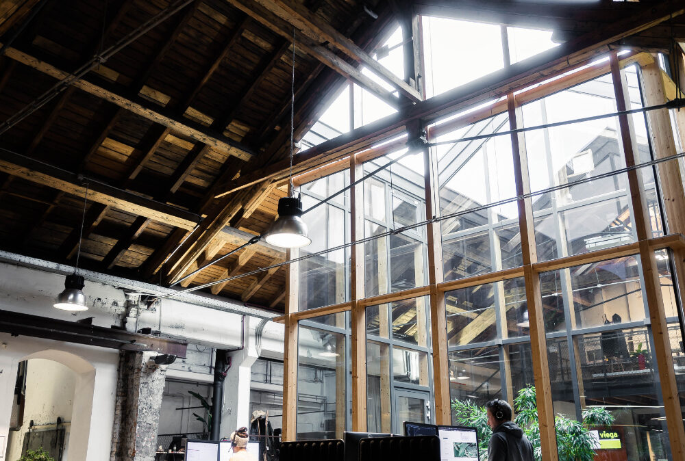 Two people are working at standing desks in a modern office with a high wooden ceiling and large glass windows.