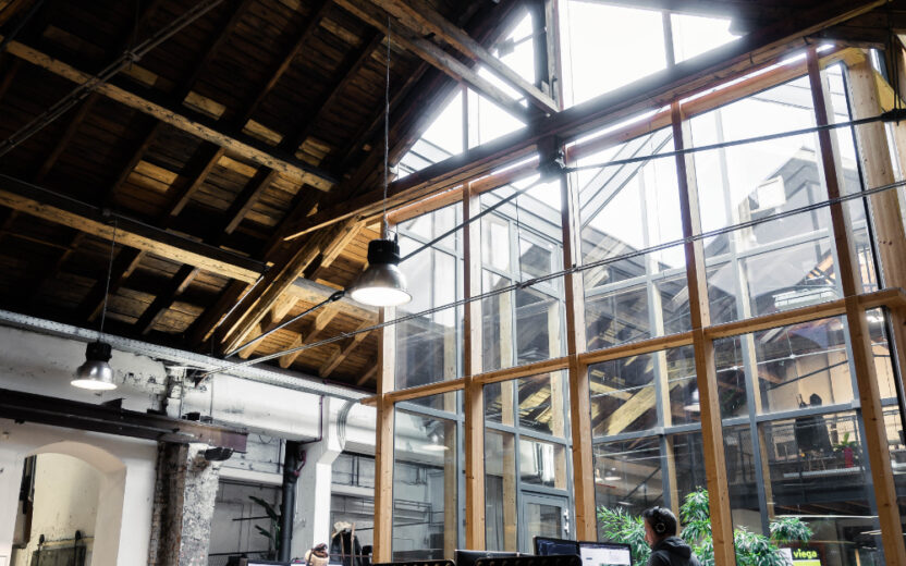 Two people are working at standing desks in a modern office with a high wooden ceiling and large glass windows.