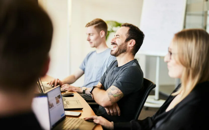 Vier Personen sitzen mit Laptops an einem Tisch und diskutieren. Eine Person lacht. Im Hintergrund ist ein Whiteboard zu sehen.
