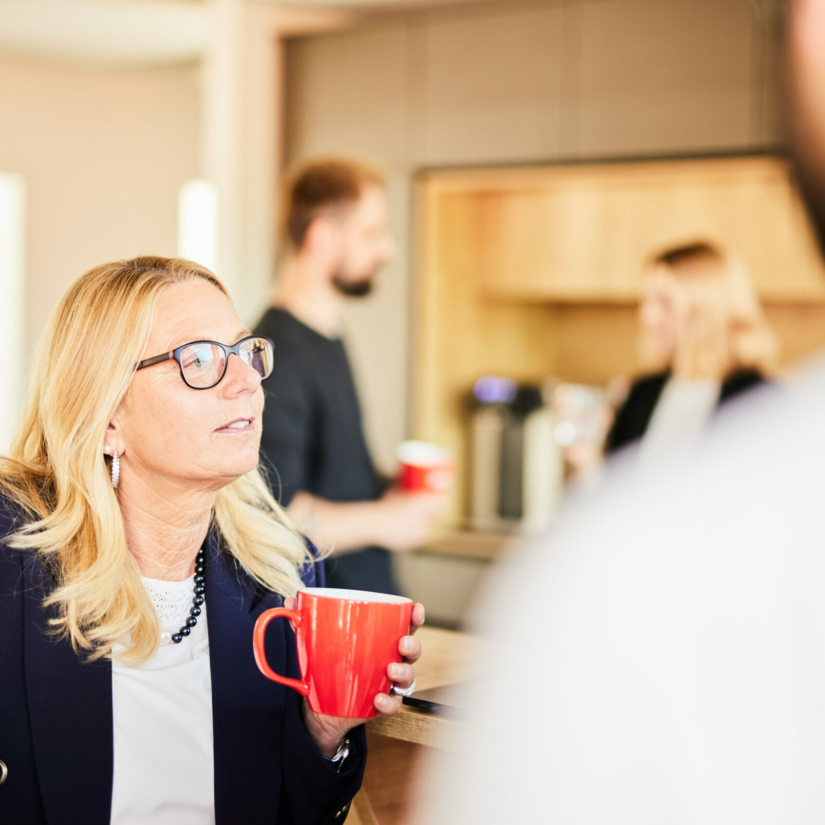 Foto von einer blonden Frau mittleren Alters, die mit einer valantic-Tasse mit ihren Kolleg*innen an der Kaffeebar ein Gespräch führt.