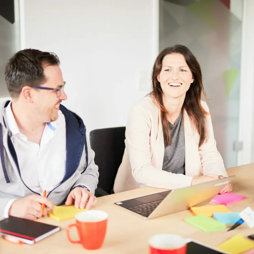 Photo of a man and woman laughing in a valantic CX client workshop. In front of them on the table are a laptop and coffee cups.