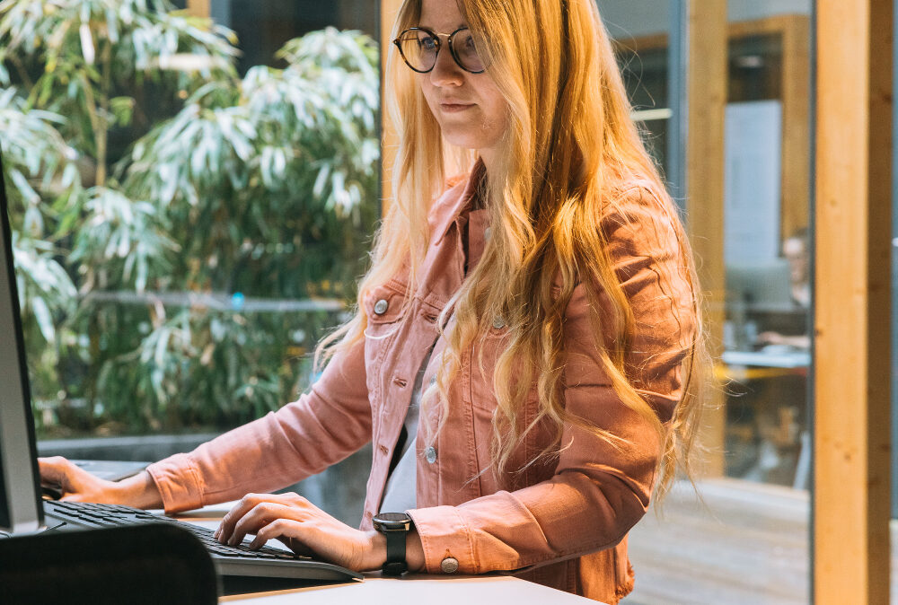 Eine Frau steht an einem Stehtisch im Büro und arbeitet konzentriert am Computer.