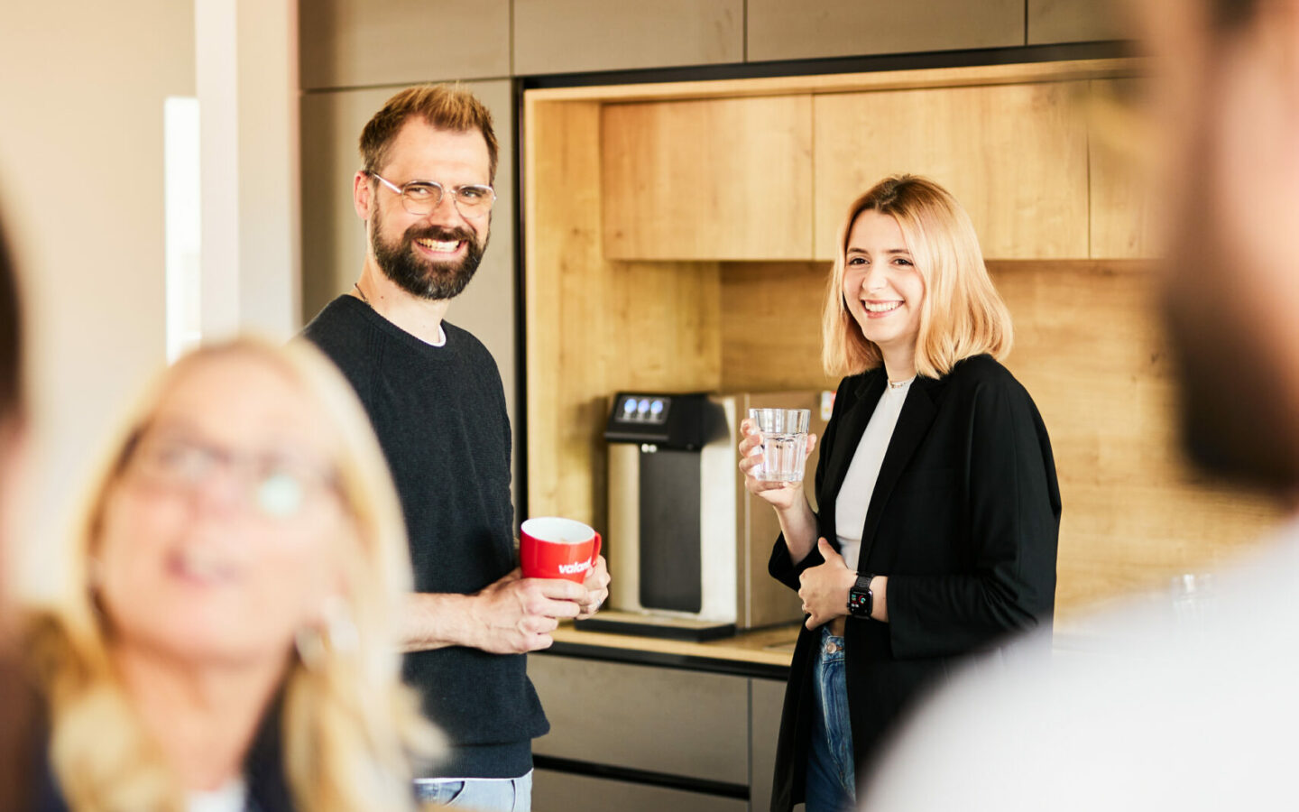 Foto von einem Mann und einer Frau, die an der Office-Bar Kaffee trinken, im Vordergrund sind unscharf andere Kolleg*innen zu erkennen.