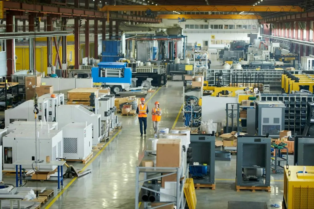 Insight into a production hall through which a man and a woman with helmet and high-visibility vest are walking