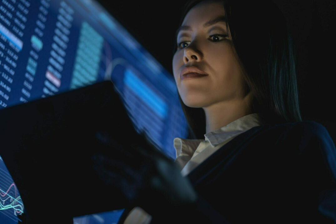 Geschäftsfrau guckt auf Tablet, im Hintergrund großer Display mit Zahlen und Verläufen