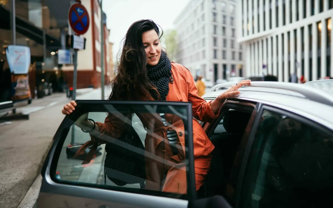 Young commuter woman catching a car ride share service in Berlin. She is smiling, wearing a fashionable coat, entering the vehicle.