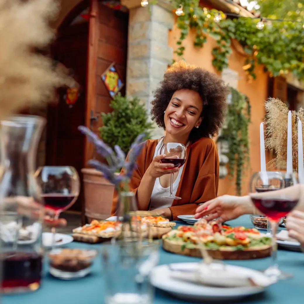 Eine Frau lächelt mit einem Glas Rotwein in der Hand an einem gedeckten Tisch im Freien, umgeben von anderen Menschen, die den Erfolg der Supply Chain Professionalisierung feiern.