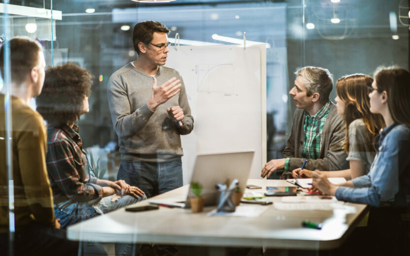 Ein Mann steht an einem Flipchart und erklärt fünf sitzenden Kollegen eine Grafik während einer Besprechung in einem modernen Büro, bei der es um LkSG-Compliance-Strategien geht.