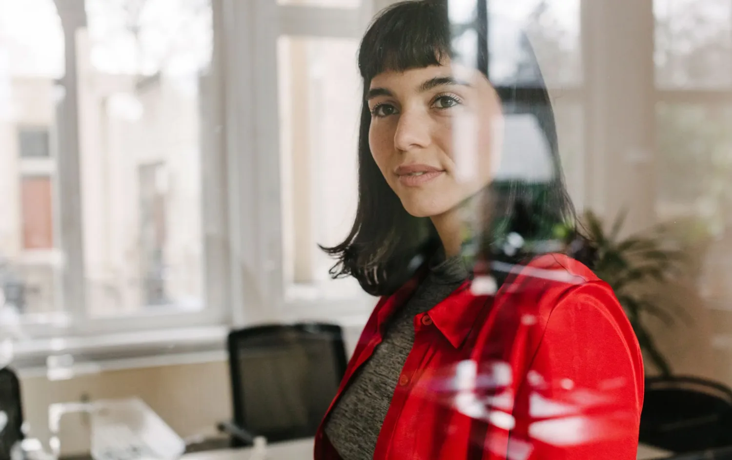 Frau mit dunklen Haaren und rotem Hemd steht in einem modernen Büro und blickt durch eine Glaswand in die Kamera - Spiegelungen als Symbol für die Digitalisierung von Versicherungen im Arbeitsumfeld.