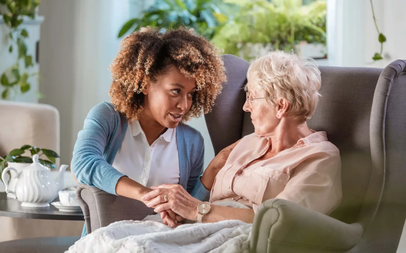 Eine jüngere Frau sitzt neben einer älteren Frau in einem Sessel, hält ihre Hand und spricht mit ihr in einem hellen, gemütlichen Raum mit Pflanzen.