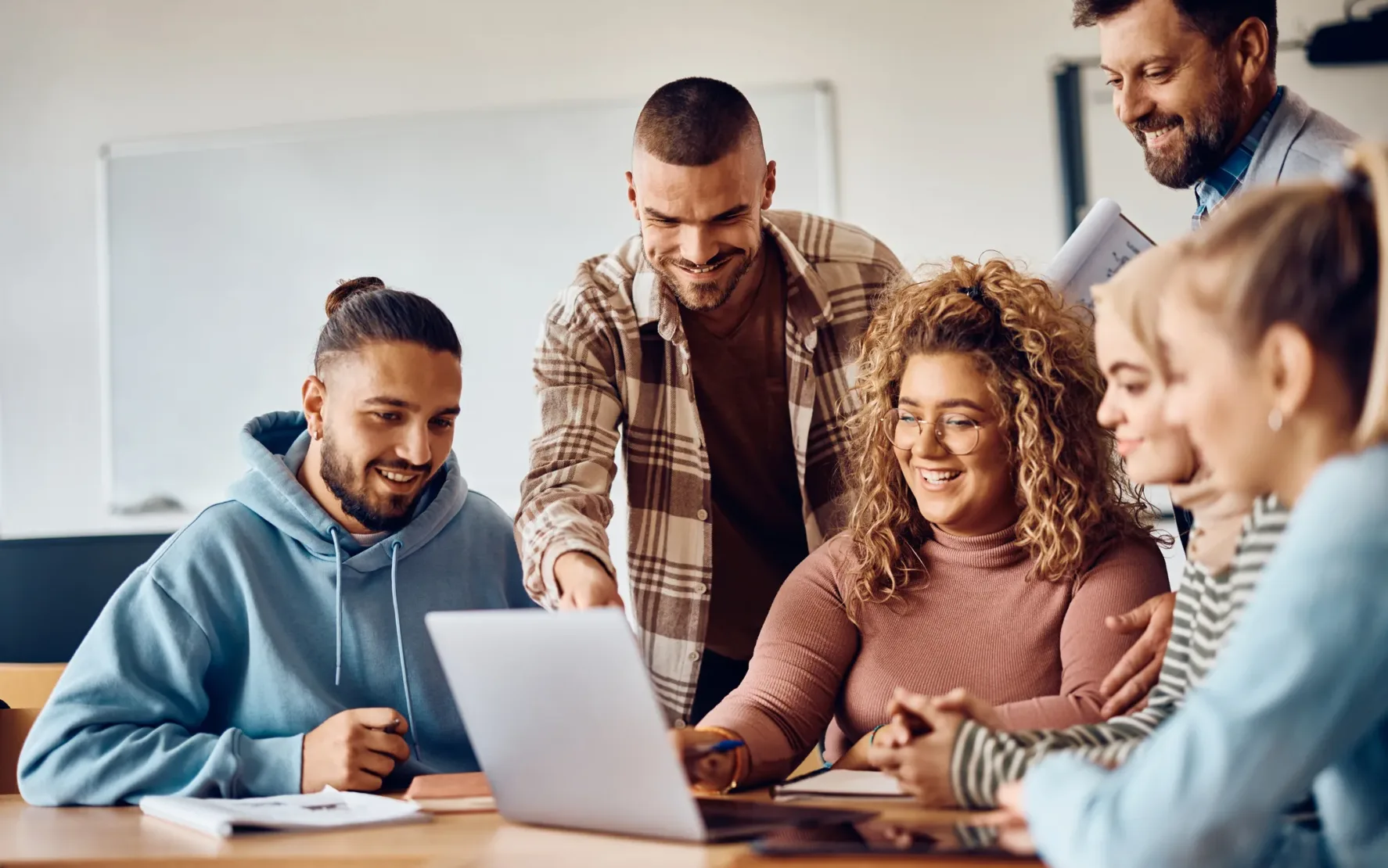 Eine Gruppe von fünf Personen, drei Männer und zwei Frauen, sitzt lächelnd um einen Tisch und arbeitet gemeinsam an einem Laptop. Sie scheinen sich während des Deutschen Low-Code-Tages in einem Klassenzimmer oder Büro zu befinden.