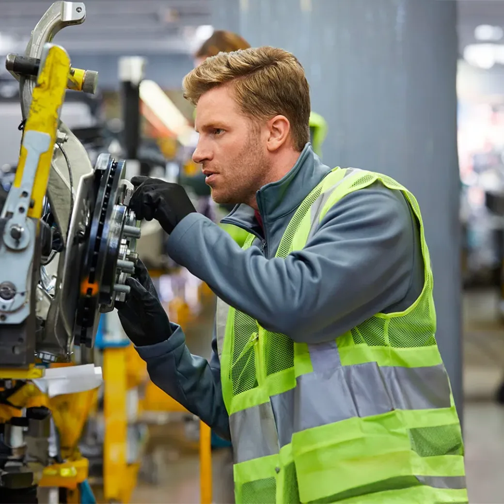 Confident mid adult engineer examining car chassis at automobile industry. Handsome male supervisor is working on car part in factory. He is wearing reflective clothing.