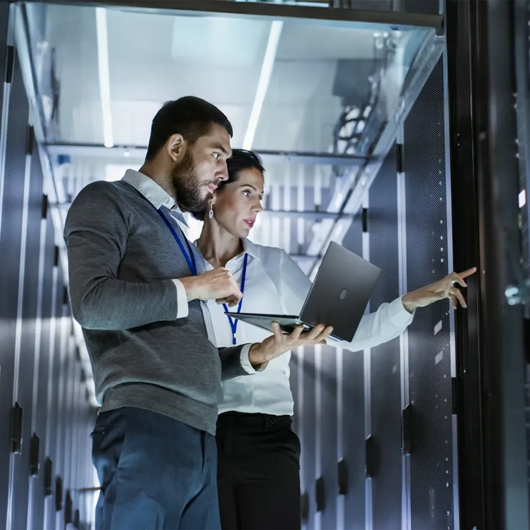 Male IT Specialist Holds Laptop and Discusses Work with Female Server Technician. They're Standing in Data Center, Rack Server Cabinet is Open. | Business Analytics