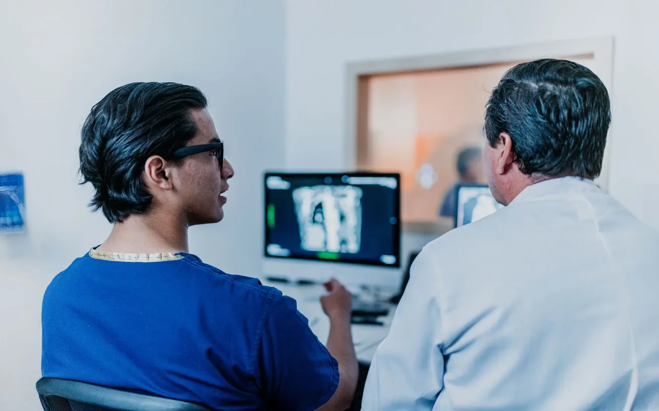 Zwei Personen in medizinischer Kleidung betrachten in einem klinischen Umfeld Diagnosebilder auf einem Computermonitor, wobei sie der Kamera den Rücken zuwenden.