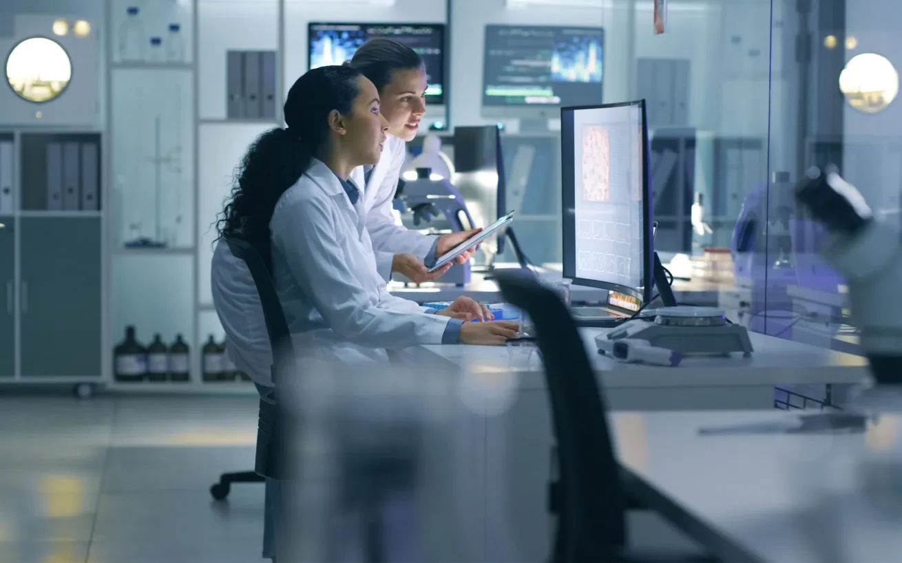 Zwei Wissenschaftler in Laborkitteln prüfen Daten auf einem Computerbildschirm in einem modernen Labor. Im Hintergrund sind wissenschaftliche Geräte und Bildschirme zu sehen.