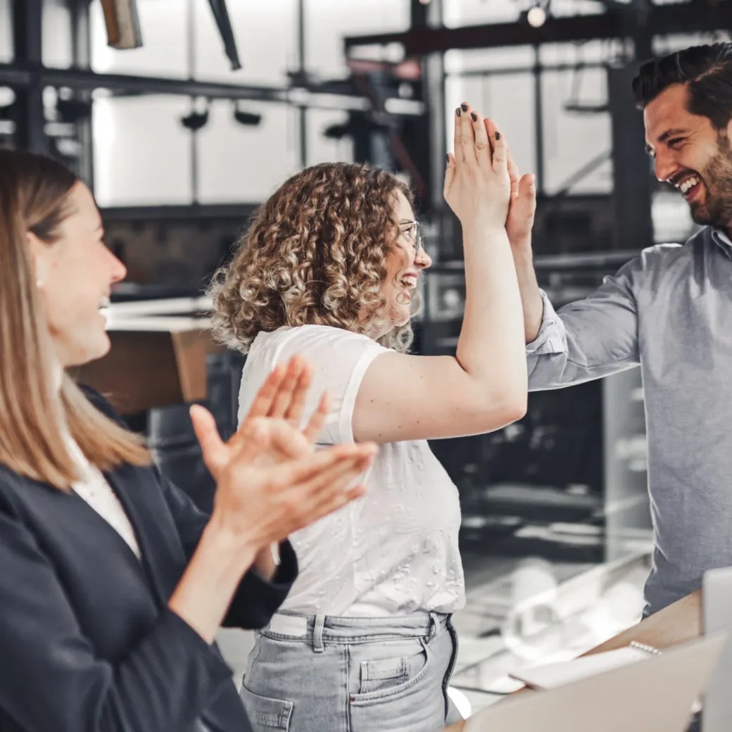 Drei Kollegen feiern Erfolge in einem modernen Büroumfeld. Zwei Personen geben sich ein High-Five, während eine andere Person in der Nähe klatscht.