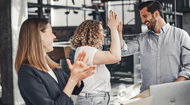 Three colleagues celebrate success in a modern office environment. Two people give each other a high-five while another person claps nearby.