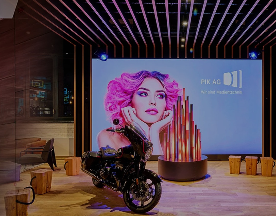 Vor einem großen Bildschirm mit "PIK Ag" ist ein Motorrad abgestellt.