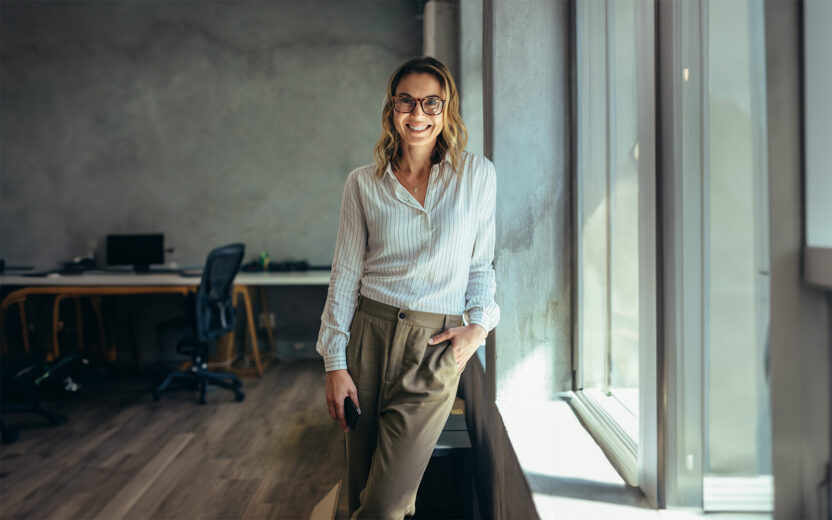 Positive businesswoman standing in office. Woman in casuals standing by window in office looking at camera.