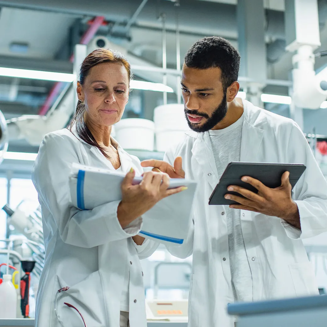Zwei Wissenschaftler in Laborkitteln prüfen Notizen und Daten auf einem Klemmbrett und einem Tablet in einer Laborumgebung.
