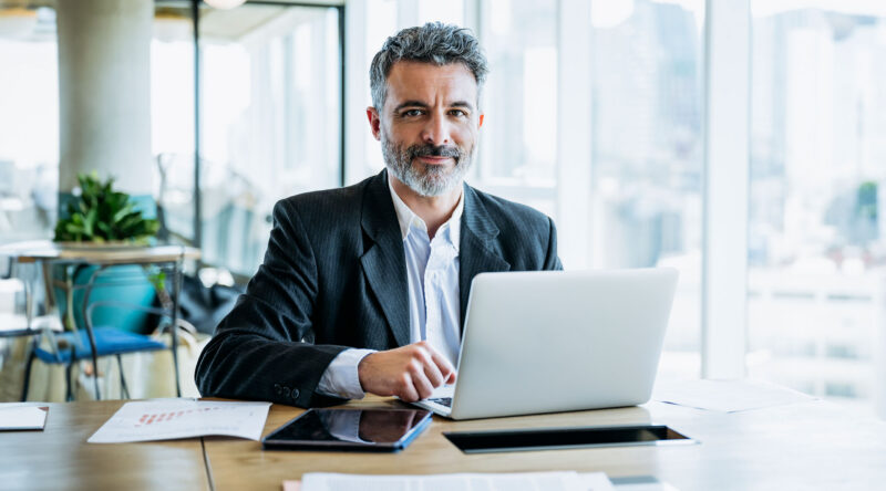 Businessman sitting at office work table next to window with view of downtown and pausing from typing on laptop to smile at camera.