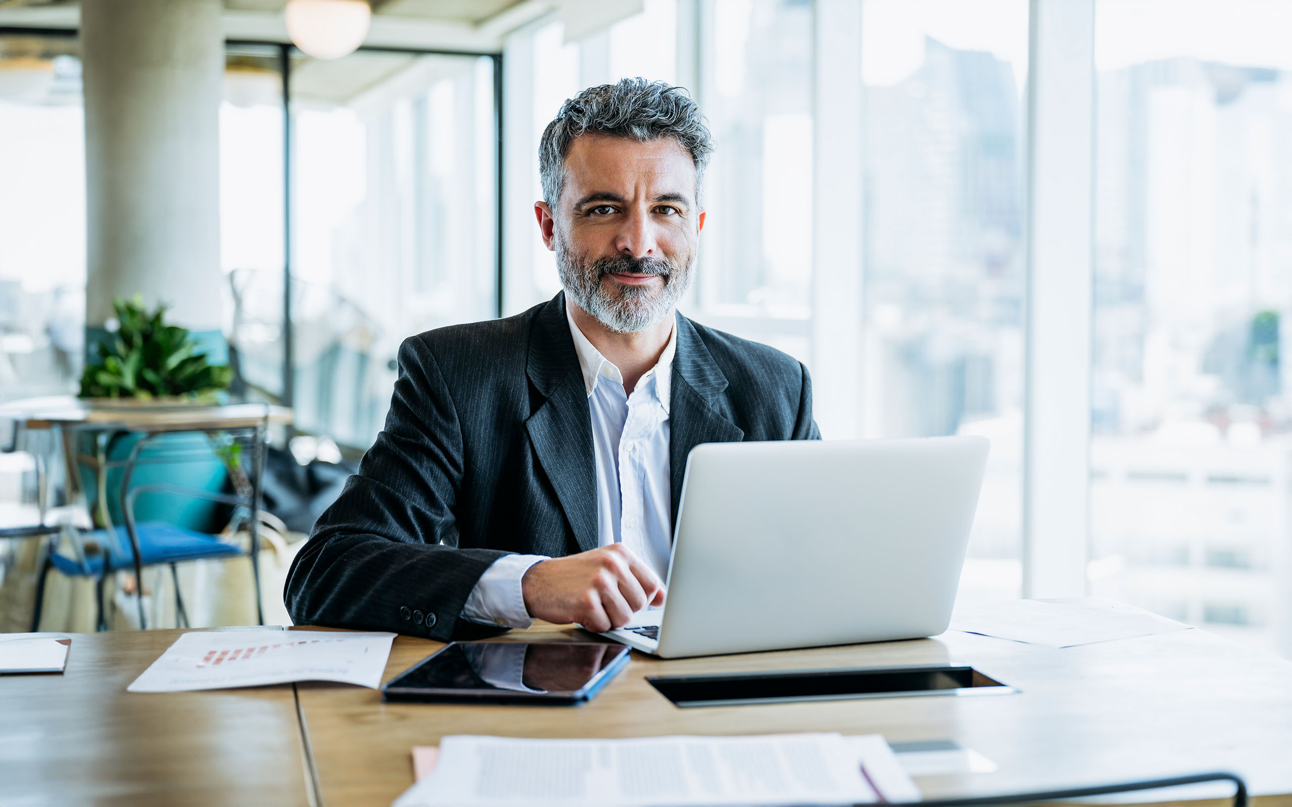 Businessman sitting at office work table next to window with view of downtown and pausing from typing on laptop to smile at camera.