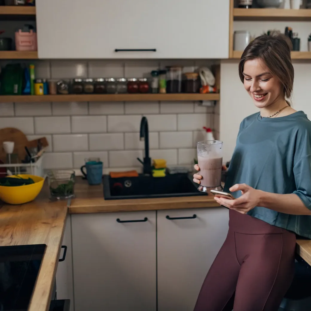 Eine Frau steht in einer Küche mit einem Smoothie in der Hand und schaut auf ihr Telefon, umgeben von frischen Produkten und Küchenutensilien. Möglicherweise überprüft sie ihren S&OP-Plan, während sie ihren Tag plant.