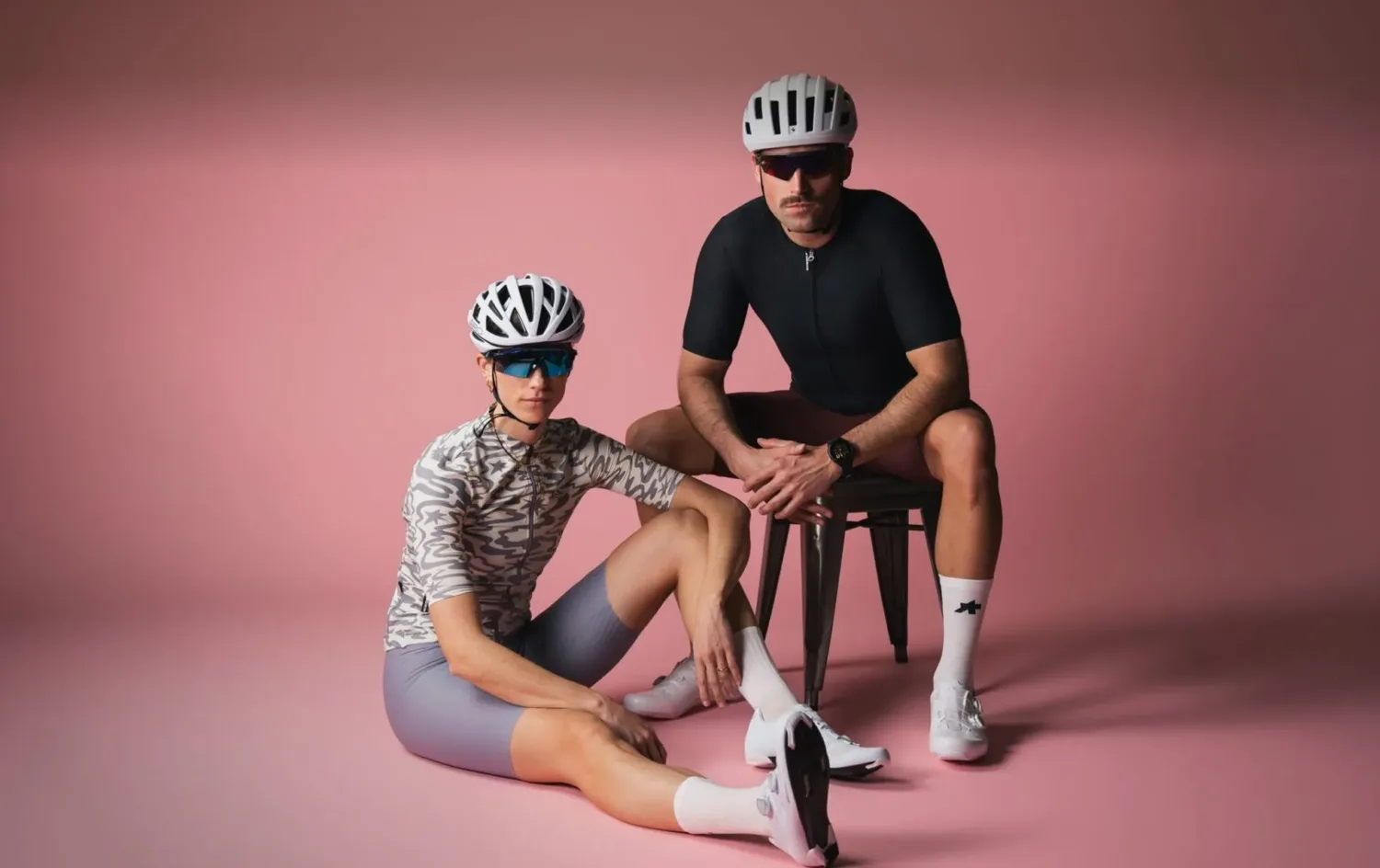 Two cyclists wearing helmets, sports glasses, and modern road cycling gear are posing in a pink studio. One person is sitting on the floor, the other on a stool. Both are wearing white cycling shoes and functional performance clothing.