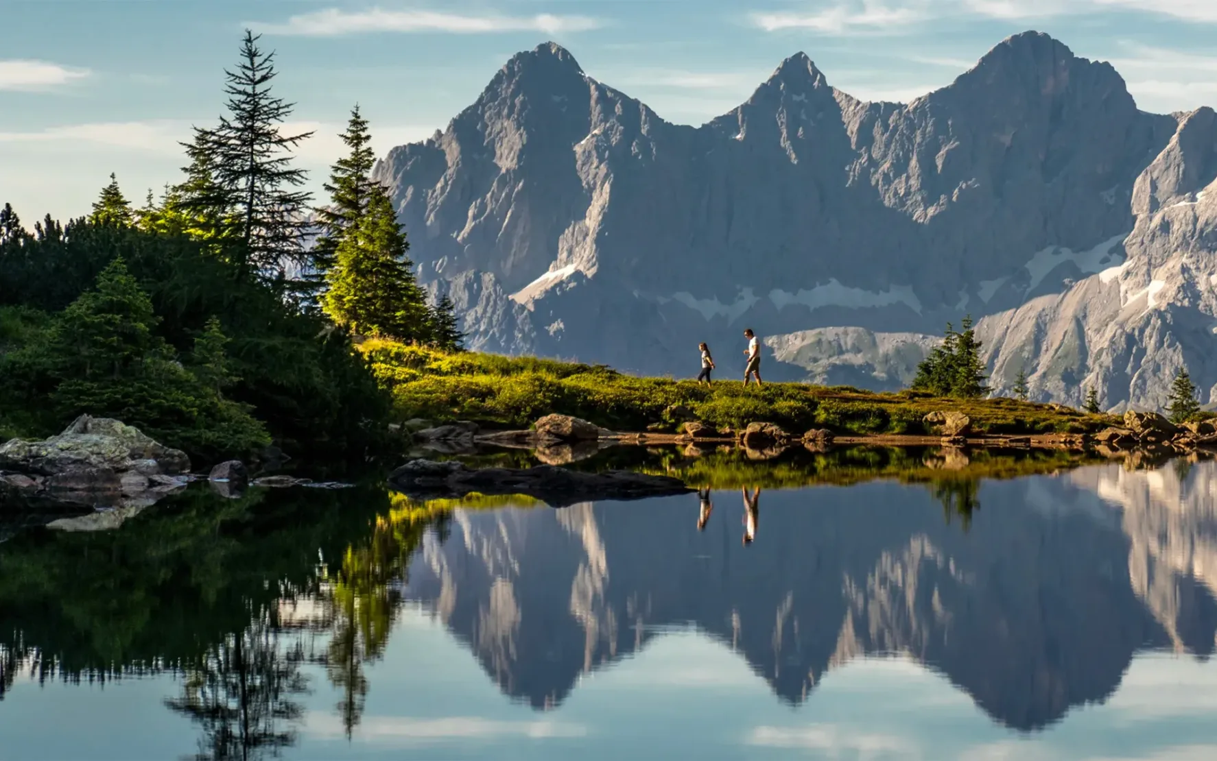 Ein Wanderer mit seinem Kind spaziert an einem sonnigen Tag entlang des Spiegelsees in der Steiermark. Im Hintergrund sieht man herrliches Bergparomana.