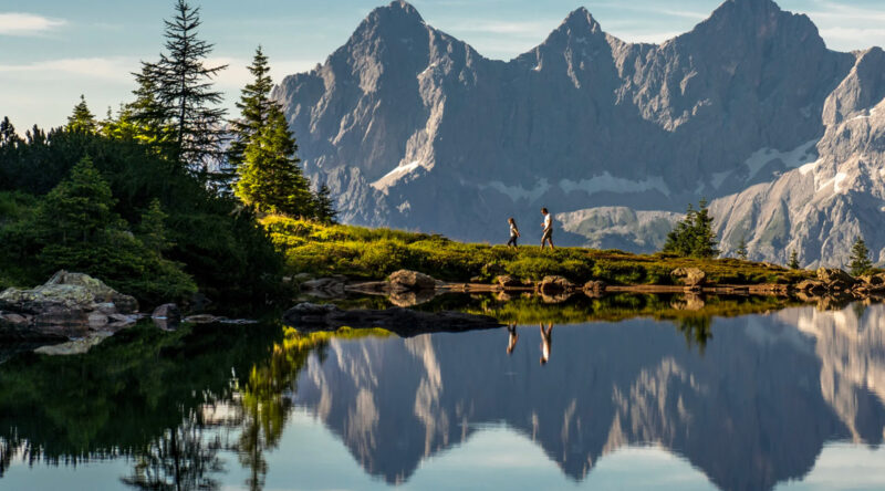Ein Wanderer mit seinem Kind spaziert an einem sonnigen Tag entlang des Spiegelsees in der Steiermark. Im Hintergrund sieht man herrliches Bergparomana.