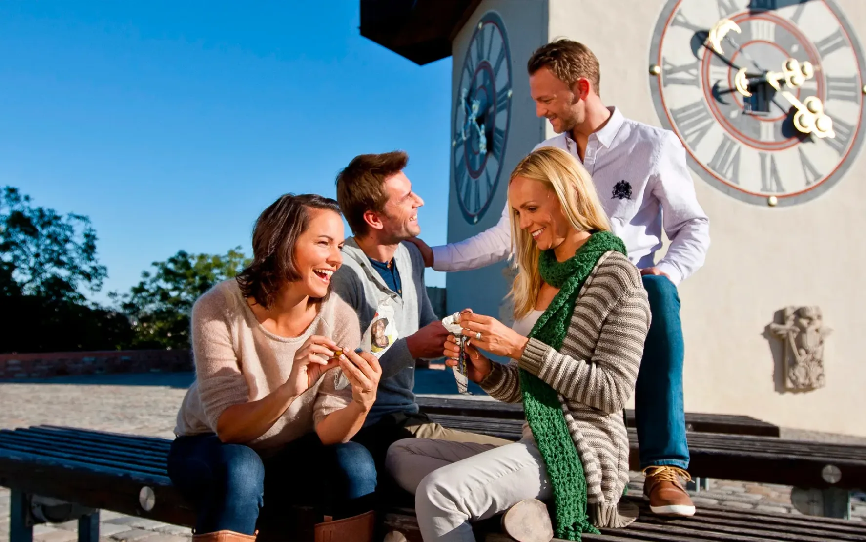 Zwei heterogene Pärchen sitzen in Graz vor dem Uhrturm in der Sonne und haben gute Laune.