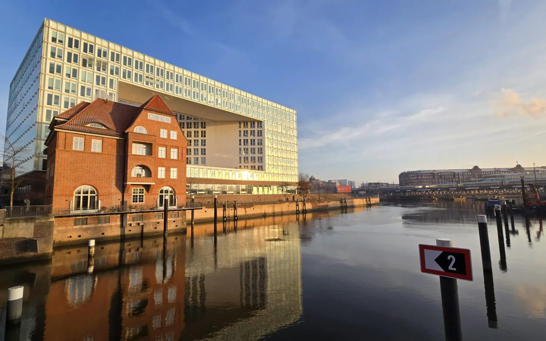 Ein modernes Glasgebäude und ein historischer roter Backsteinbau stehen an einem ruhigen Kanal unter strahlend blauem Himmel und spiegeln sich im Wasser.