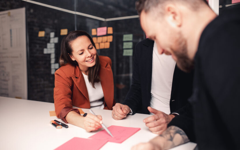 Drei Personen sitzen mit Haftnotizen und Stiften an einem Tisch und diskutieren über die NIS-2-Richtlinie. Eine Person schreibt lächelnd auf rosa Papier, die anderen beobachten aufmerksam.