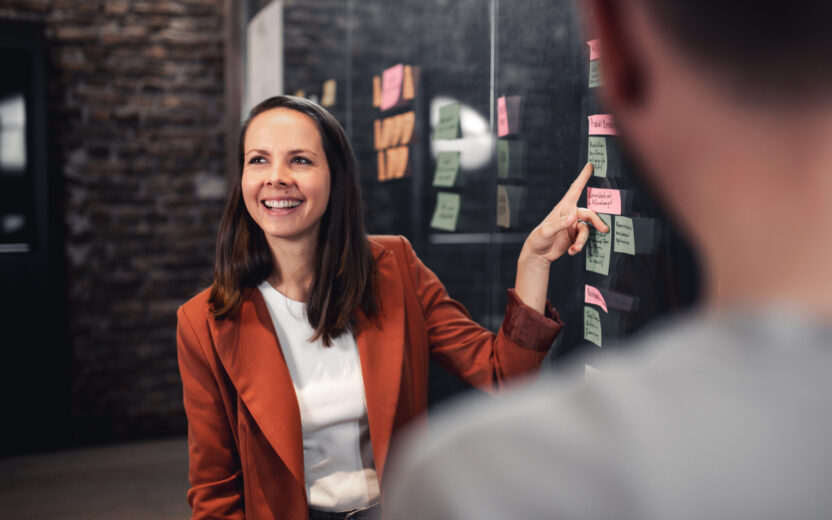 Eine lächelnde Frau mit einem orangen Blazer zeigt mit dem Finger auf ein Post-It.