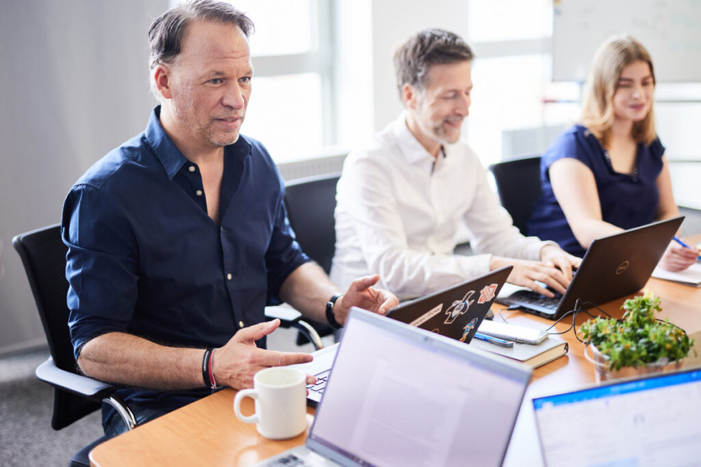 Drei Personen sitzen mit Laptops an einem Konferenztisch und diskutieren.