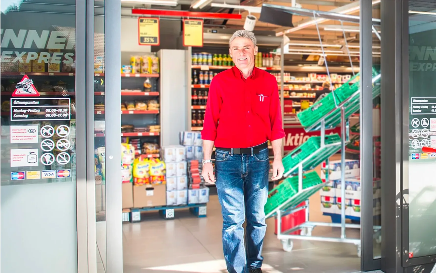 Ein Mitarbeiter steht vor dem Eingang einer Denner-Filiale und lächelt in die Kamera.