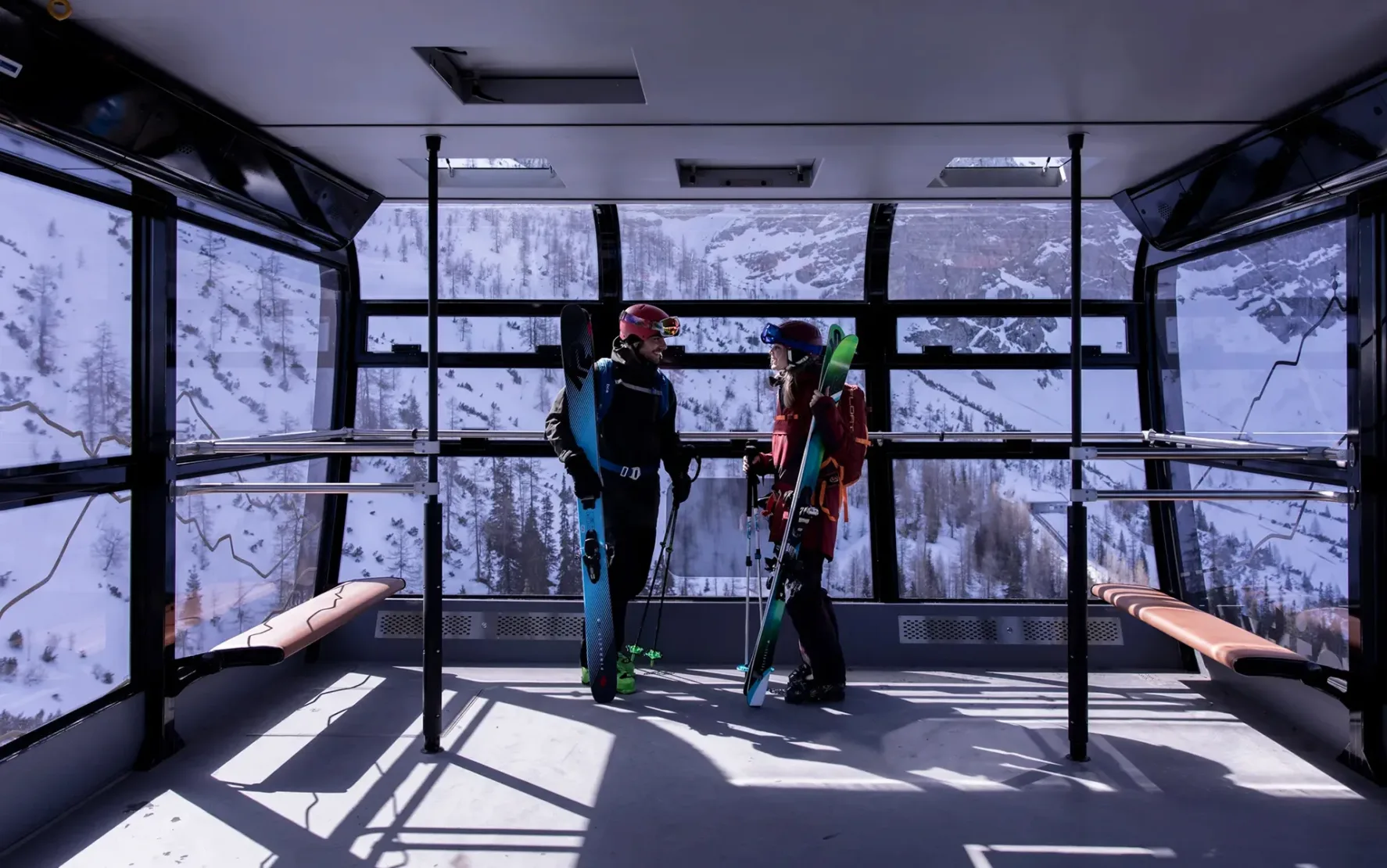 Two skiers with their gear standing in a modern, almost empty cable car of the Bayerische Zugspitzbahn, surrounded by snow-covered mountains in the background.