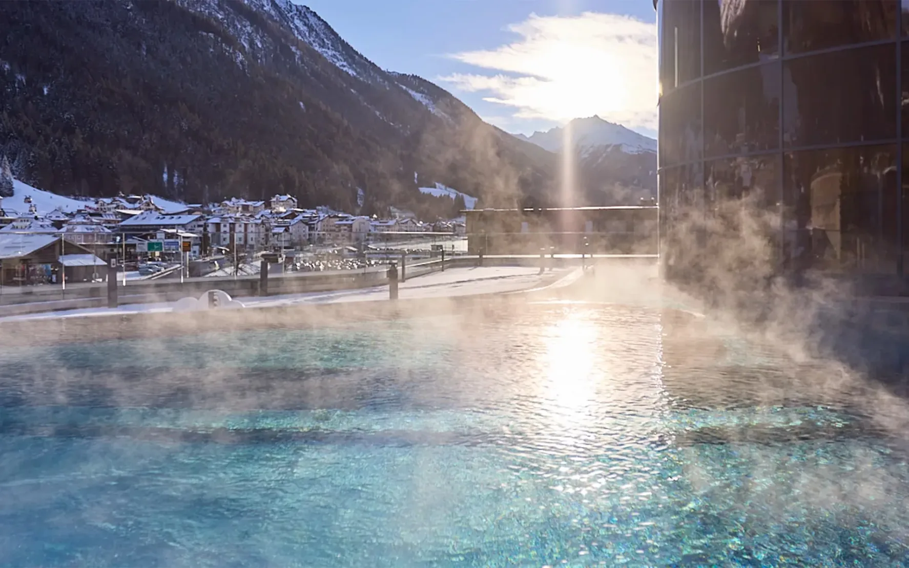 Außenbecken der Silvretta Therme Ischgl mit Blick auf das winterliche Ischgl und die umliegende Berglandschaft bei Sonnenaufgang.